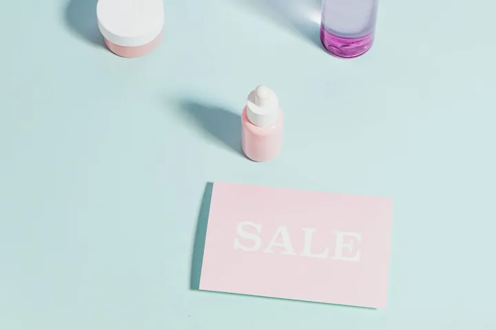 Pastel skincare bottles and a small card reading SALE on a mint and lilac background