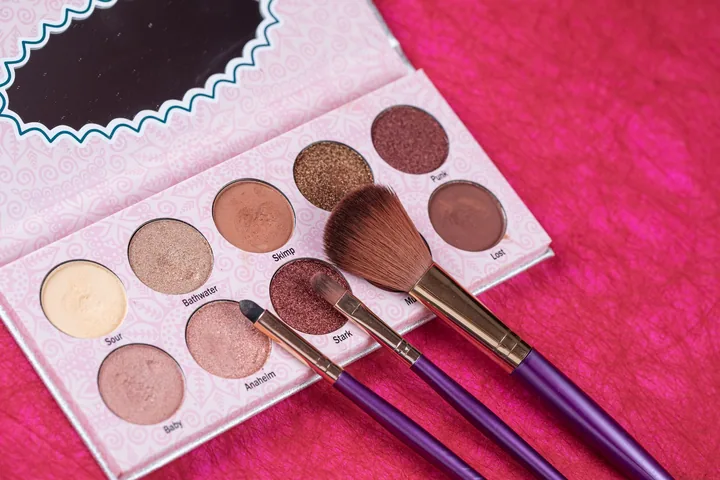 A neutral eyeshadow palette and two makeup brushes on bright pink fabric
