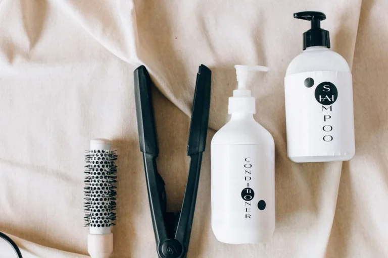 A flat iron, round brush and unbranded hair-care bottles on a cream blanket
