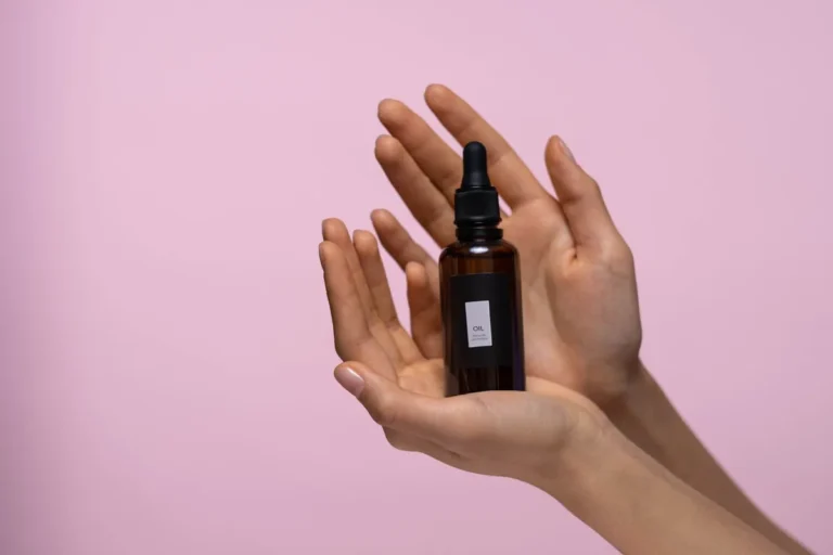 Two cupped hands holding a small amber serum dropper bottle against a soft pink backdrop