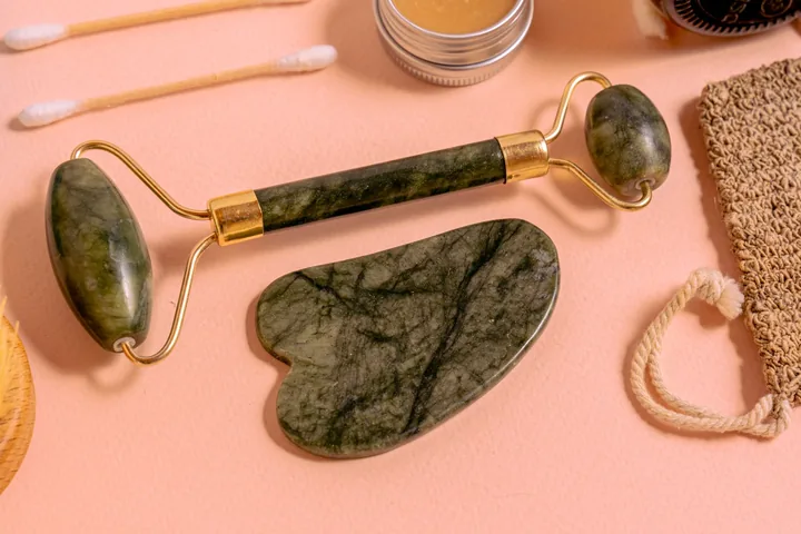 A jade facial roller, gua sha and small skincare jars on a peach-pink surface