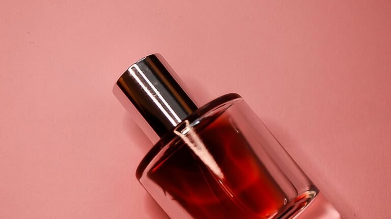 A glass perfume bottle filled with deep red-pink fragrance lying on a pastel pink surface