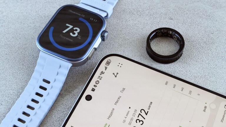 A smartwatch with a pale strap, a black smart ring and a phone showing step data on a grey surface